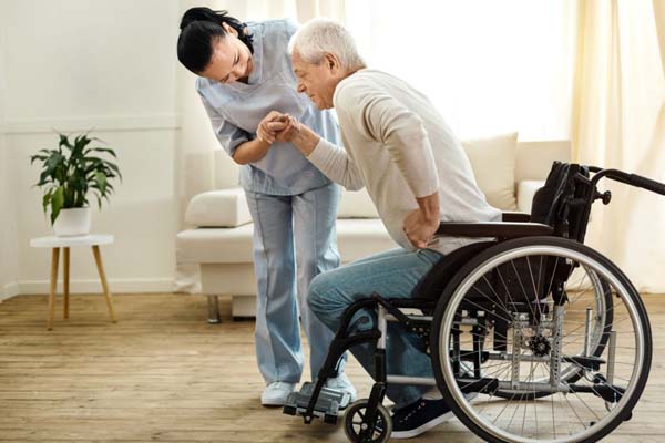 Carer supporting elderly man in wheelchair with a physical disability stand up