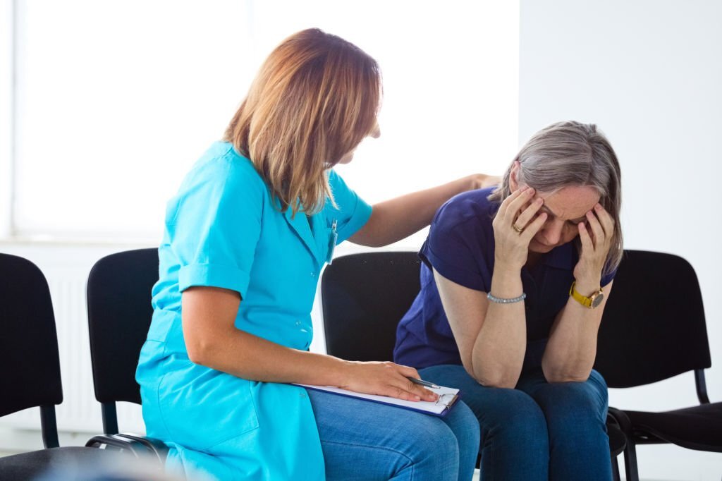 Nurse consoling an elderly lady. They are sitting on chairs in waiting room in the hospital.